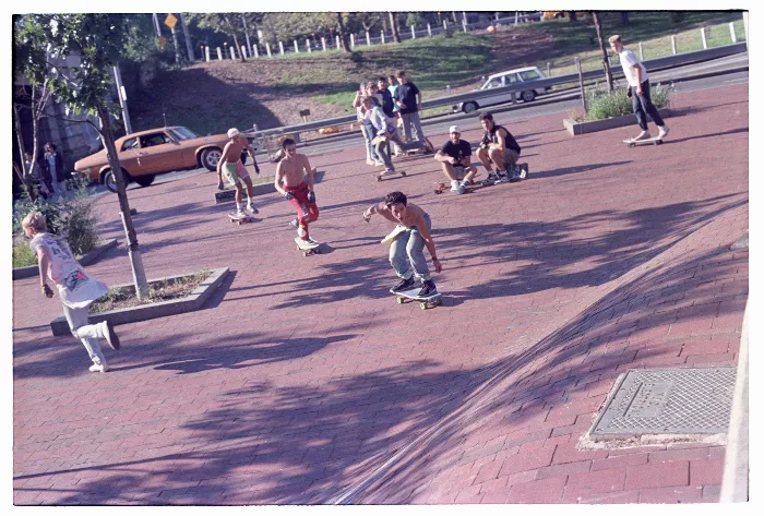 A black and white photo shows a side view of a skateboarder mid-air, ollieing over a wall, with spectators on the side