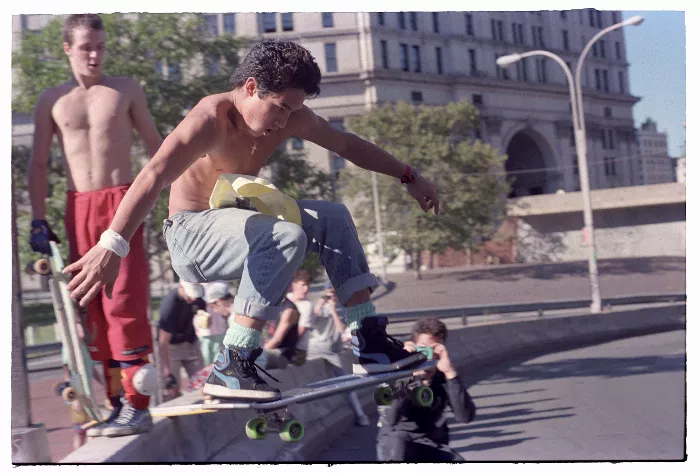 A black and white photo shows a skateboarder ollieing over a wall, with spectators watching in the background