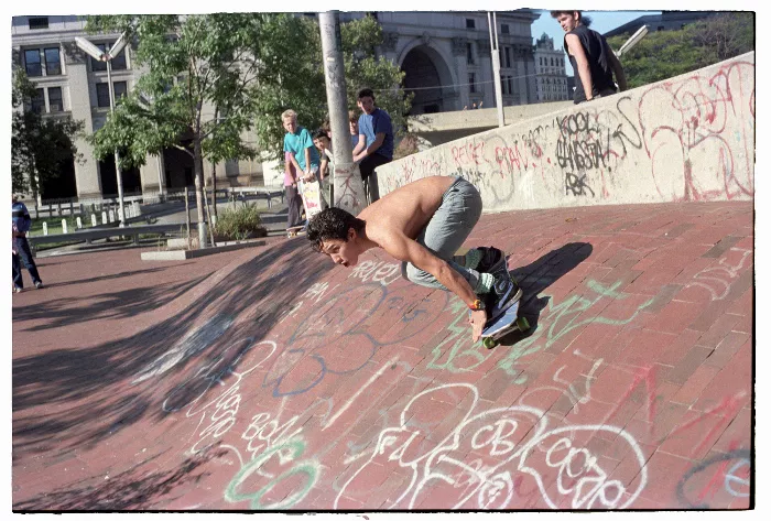 A black and white photo shows a skateboarder performing a trick on a ledge with another skateboarder in the background