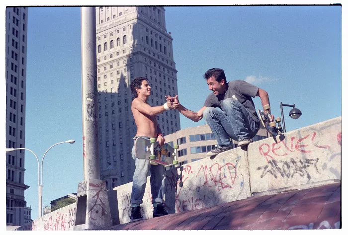 A black and white photo shows a skateboarder performing a trick on a ledge with people in the background