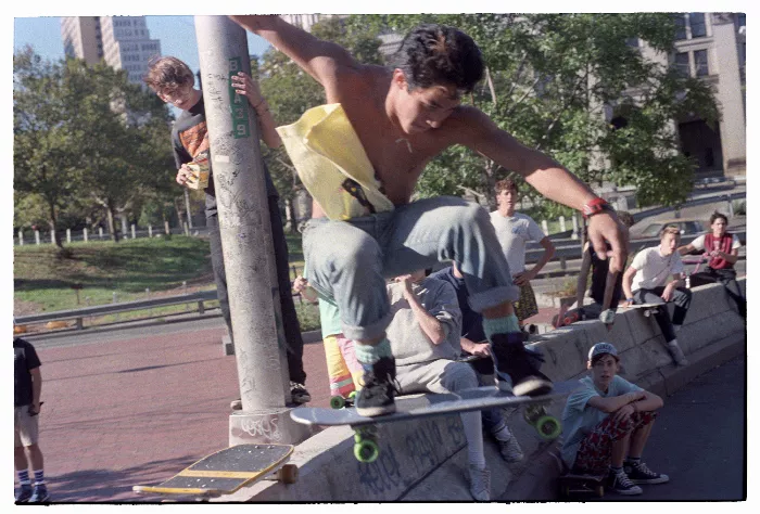 A color photo shows a group of people watching a skateboarder jumping over a high wall