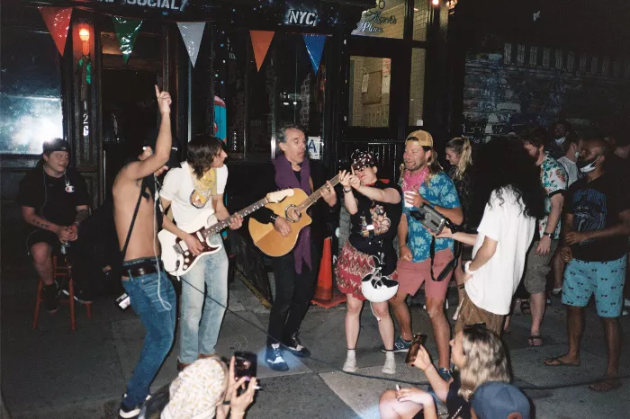 A group of people gathered closely together at night, some without masks, with a few holding drinks.