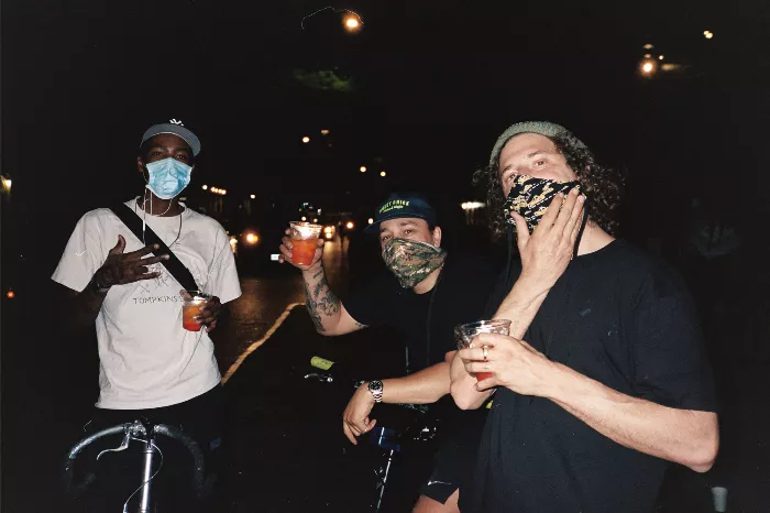 A group of people sit on a curb enjoying drinks and snacks, some on bikes, celebrating a birthday on the street.