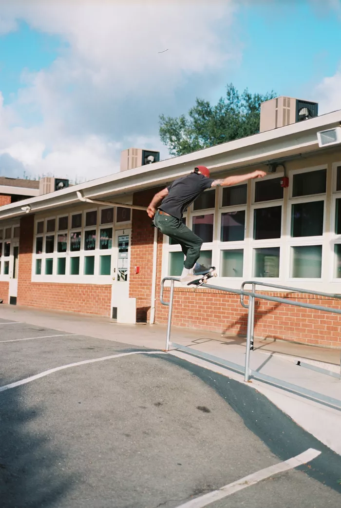 A longboarder in mid-air, performing a trick on a ramp in a skate park, with other skaters visible in the background.