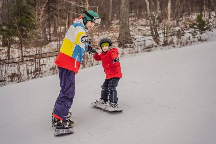 A man in a red jacket is teaching a child wearing blue to snowboard, holding their hands on a snowy slope.