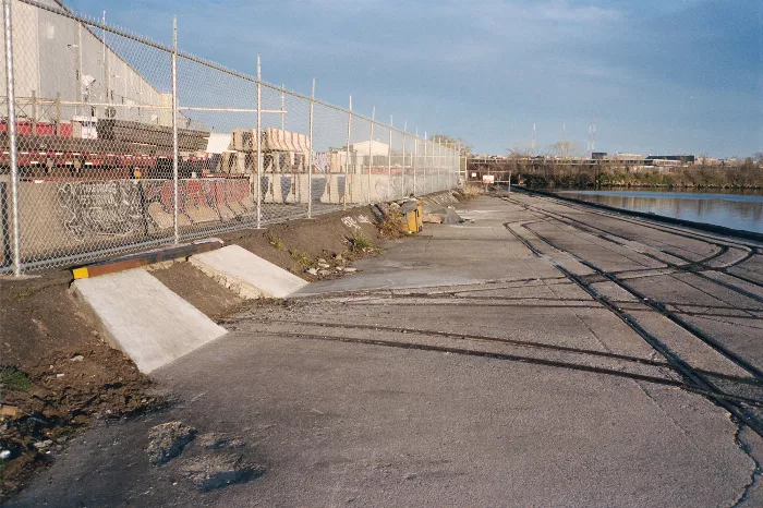 A skateboarder performs a trick on a makeshift ramp near a set of train tracks, with urban debris and uneven ground surrounding the area.