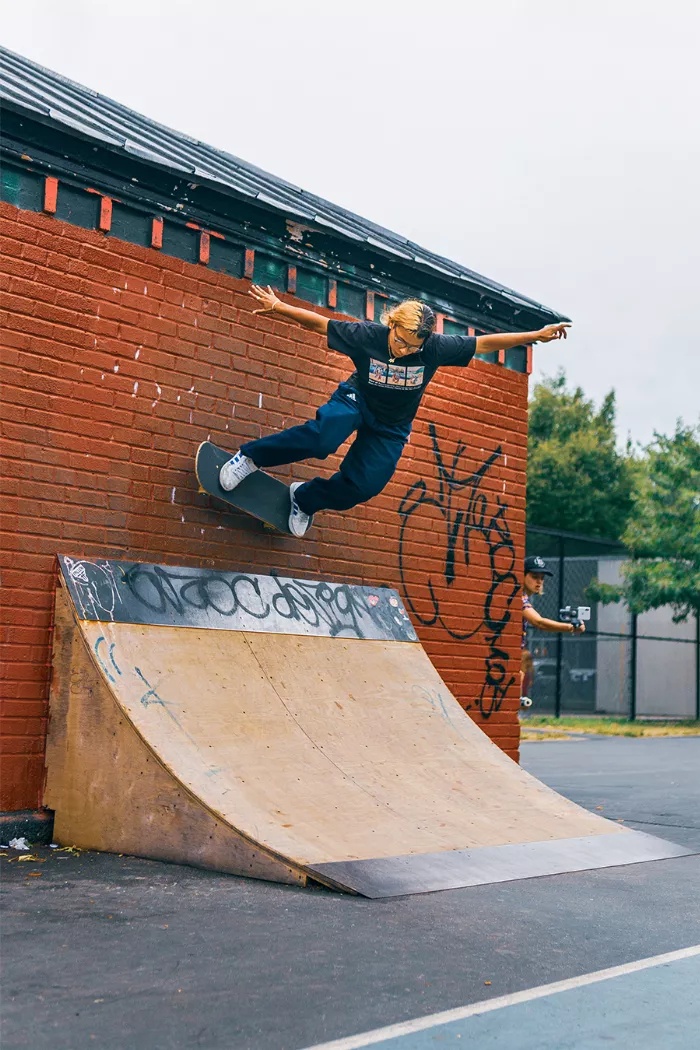 A skater performing a trick on a concrete ramp at Blue Park, with various other obstacles and people in the background.