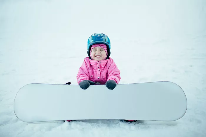 A young child in a blue snowsuit is smiling while snowboarding down a gentle slope.