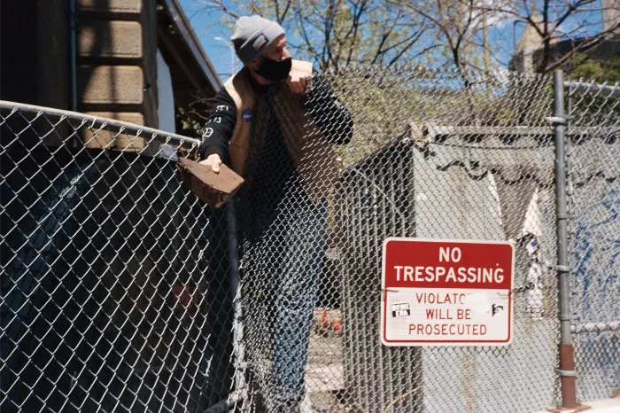 Andrew Murrell stands next to a partially demolished brick wall at the Brooklyn Banks, holding a brick in his hand, with a metal fence in the background.