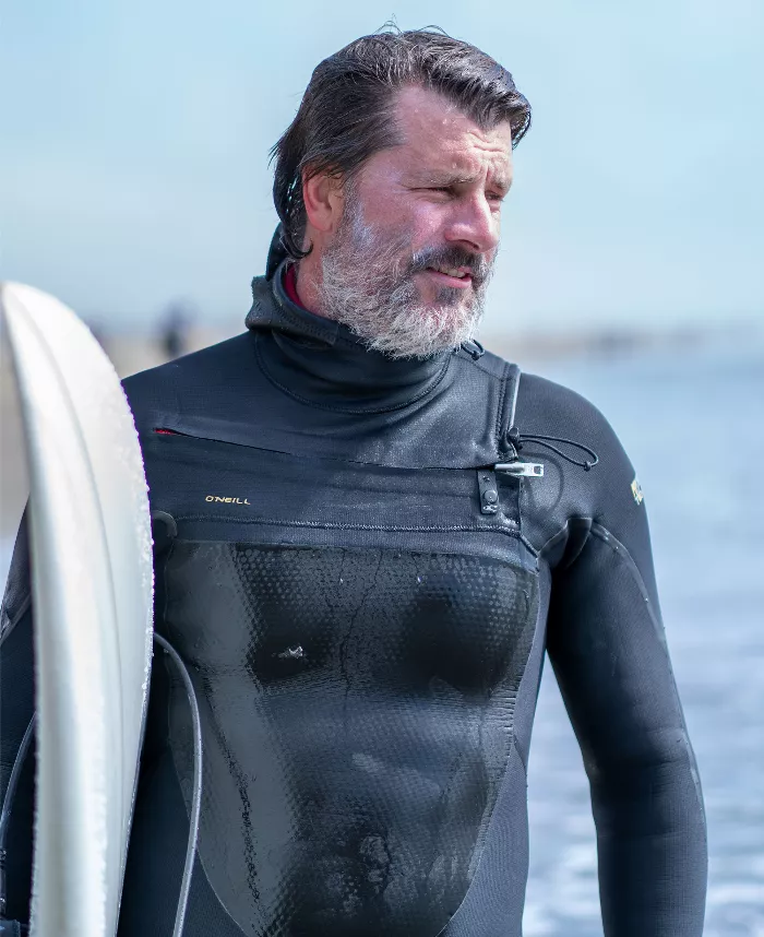 Rob Welsh, pictured from the waist up, holding a surfboard, with a sunny backdrop behind him.