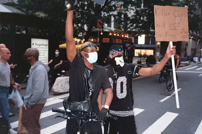 Two individuals stand on a city street with graffiti and signs in the background, one in a fashionable outfit and the other in a simple black t-shirt.